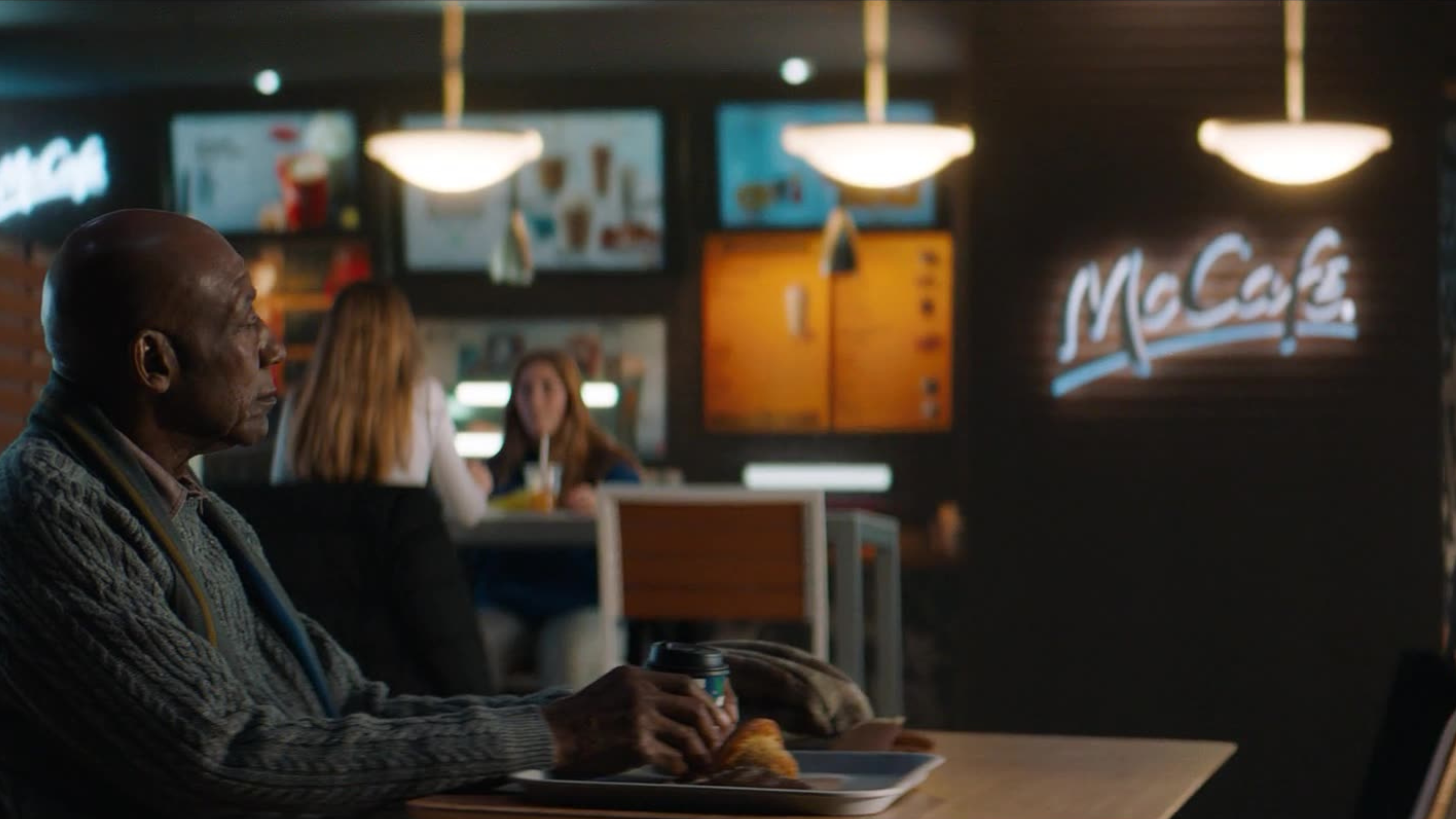 An elderly man sits quietly at a McDonald's McCafé, holding a coffee cup with a pastry on his tray.