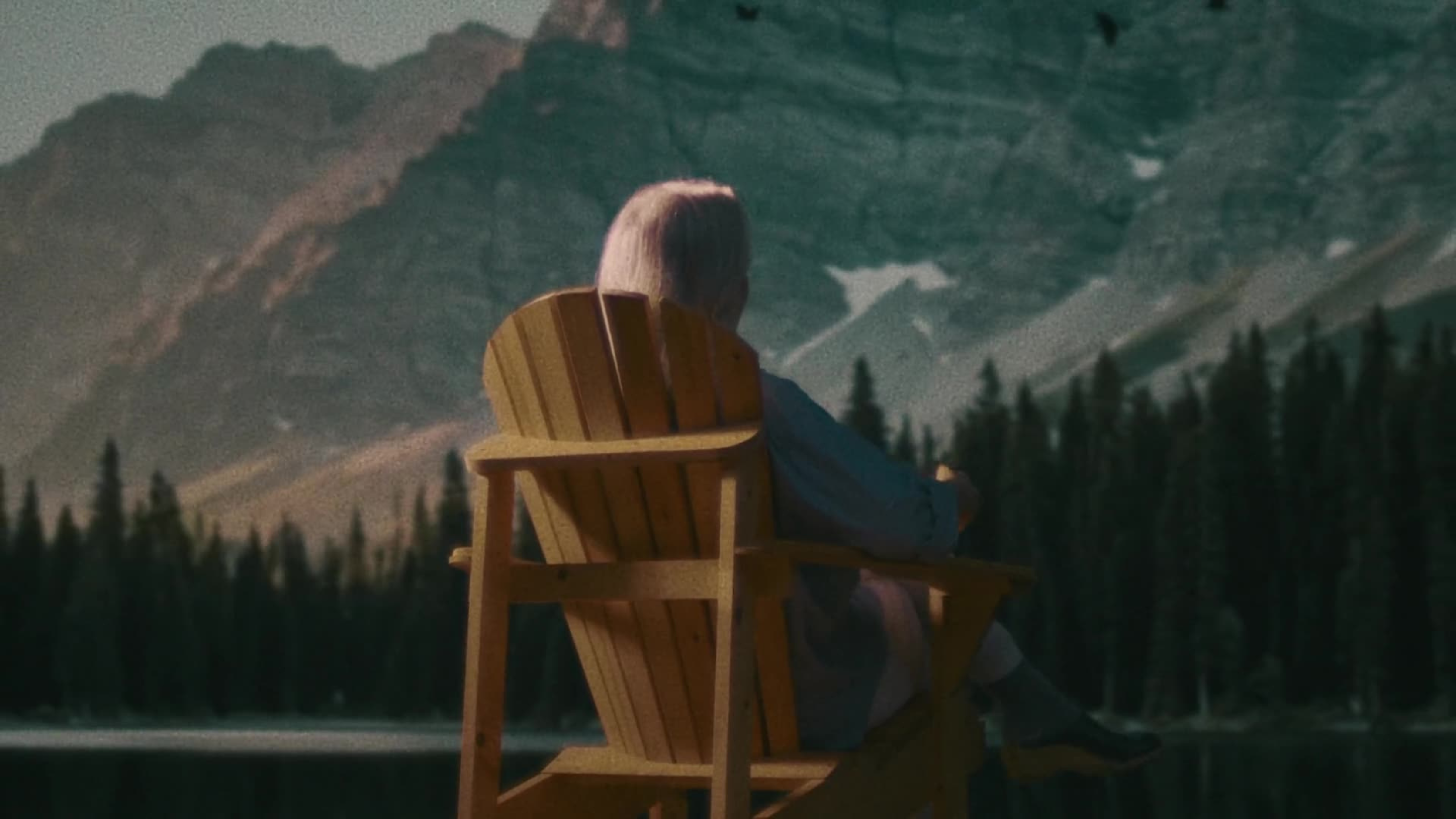 An elderly person sits alone in a wooden Adirondack chair gazing at a dramatic mountain lake landscape.