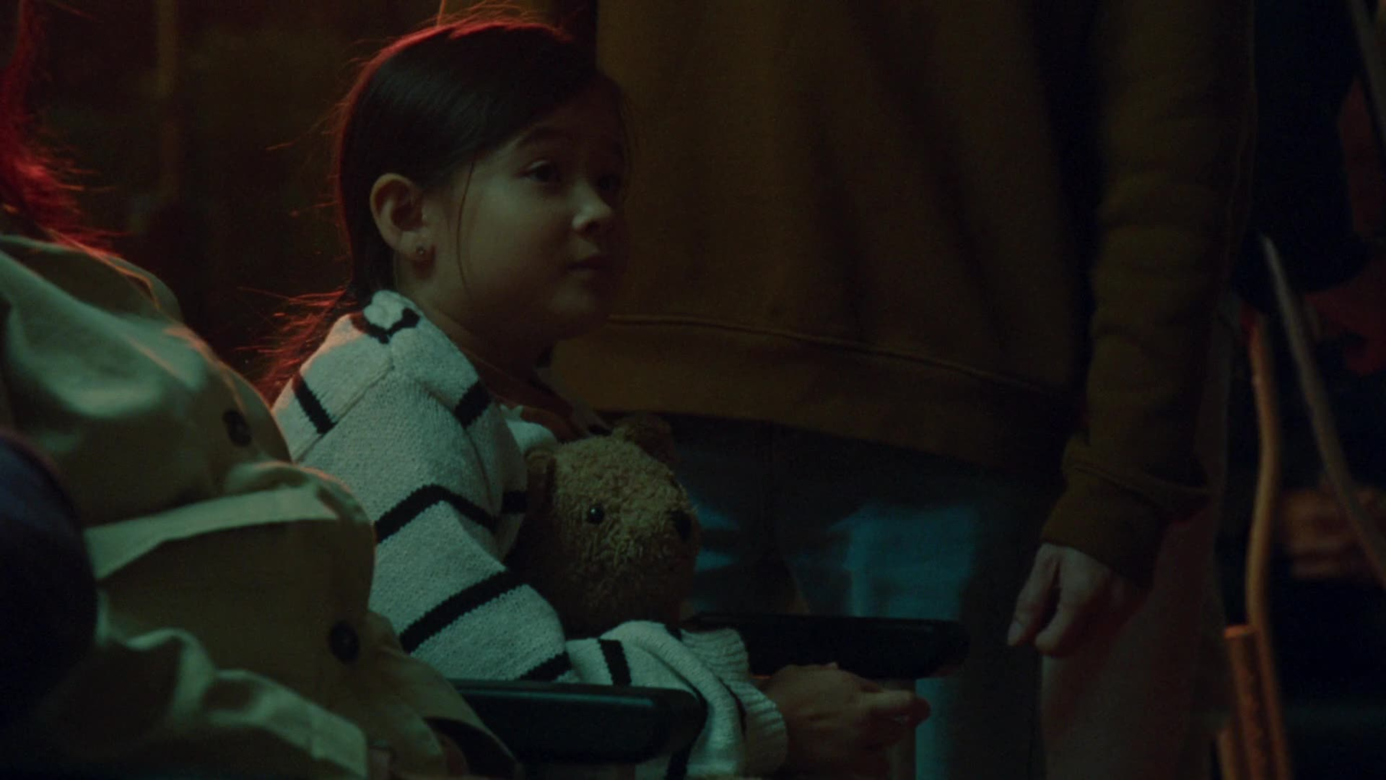 A young girl clutches a teddy bear in a dimly lit, moody scene bathed in red and green light.