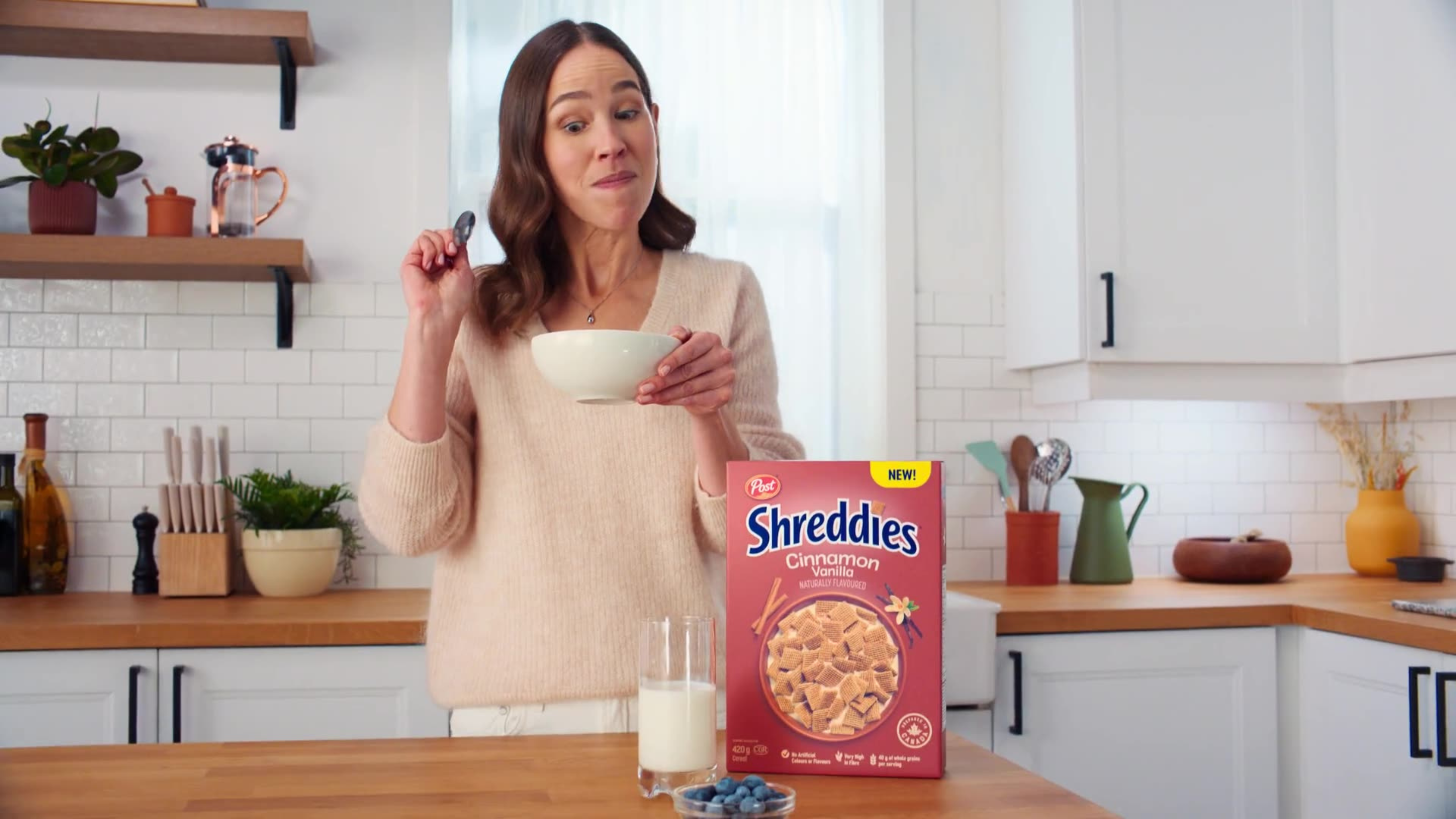 A woman holds a cereal bowl in a bright kitchen next to a box of new Post Shreddies Cinnamon Vanilla.
