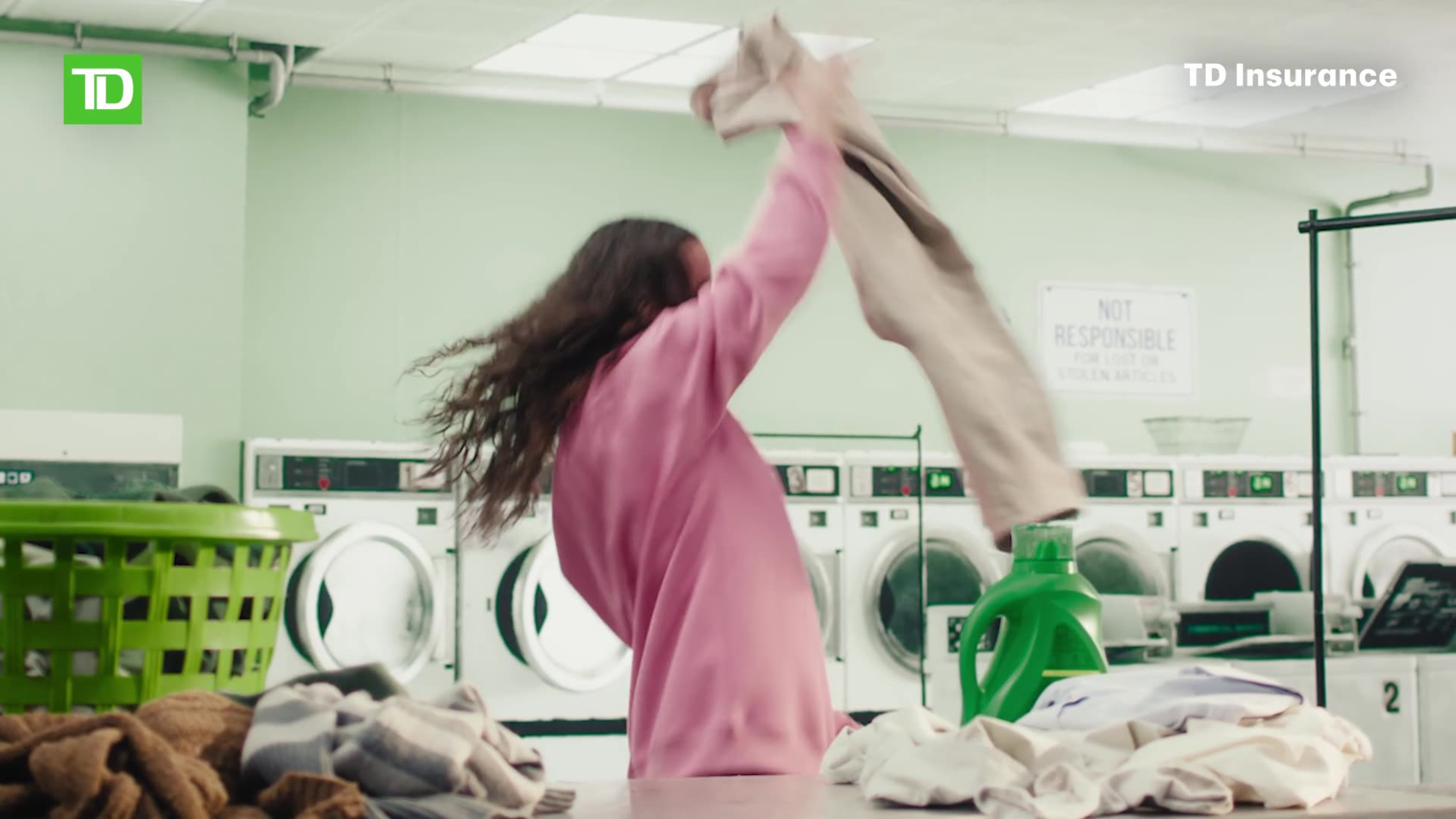 A woman in a pink hoodie twirls laundry dramatically in a laundromat in a TD Insurance ad.