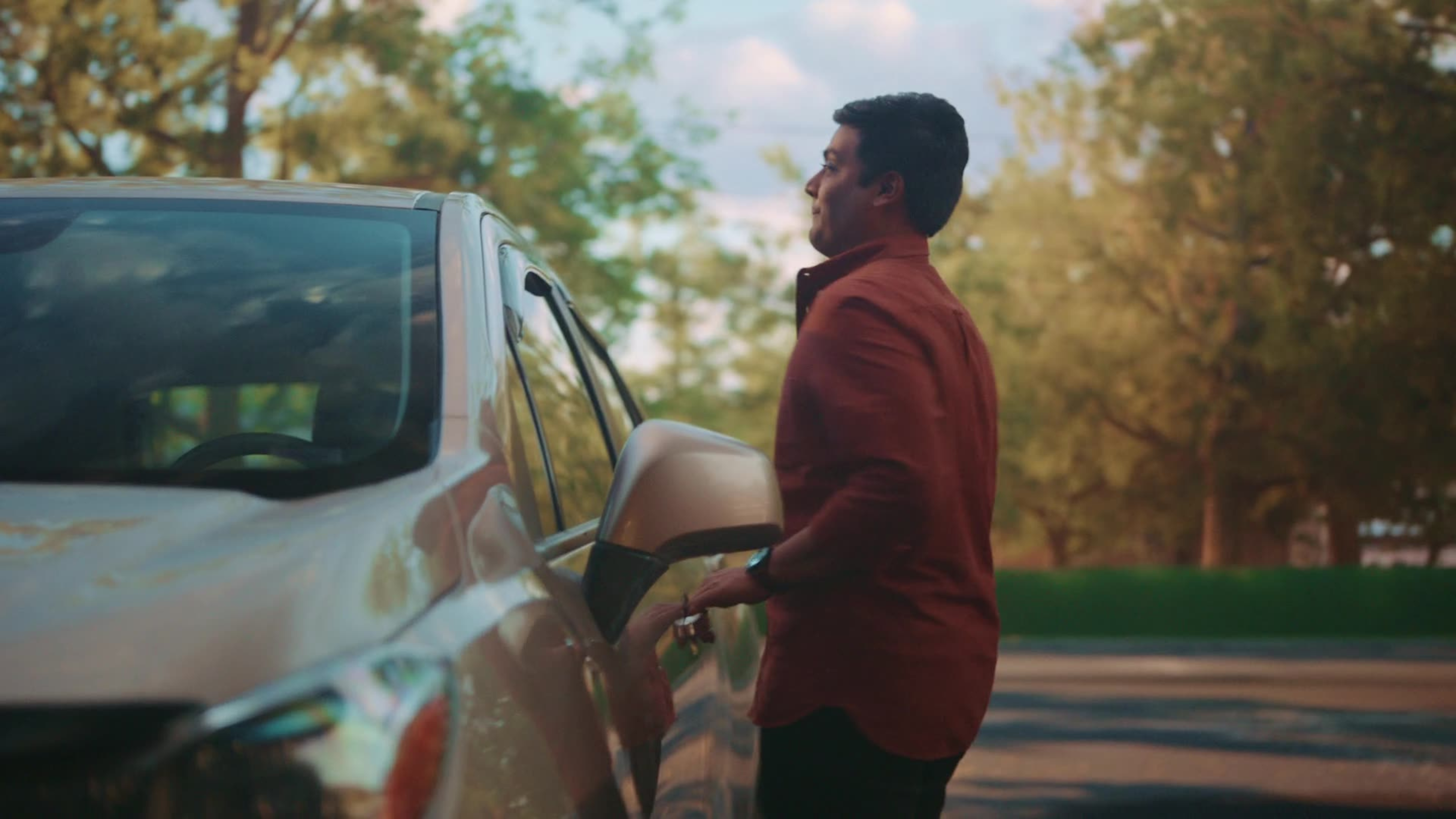 A man in a red shirt pauses thoughtfully while opening the door of a silver vehicle on a tree-lined street.
