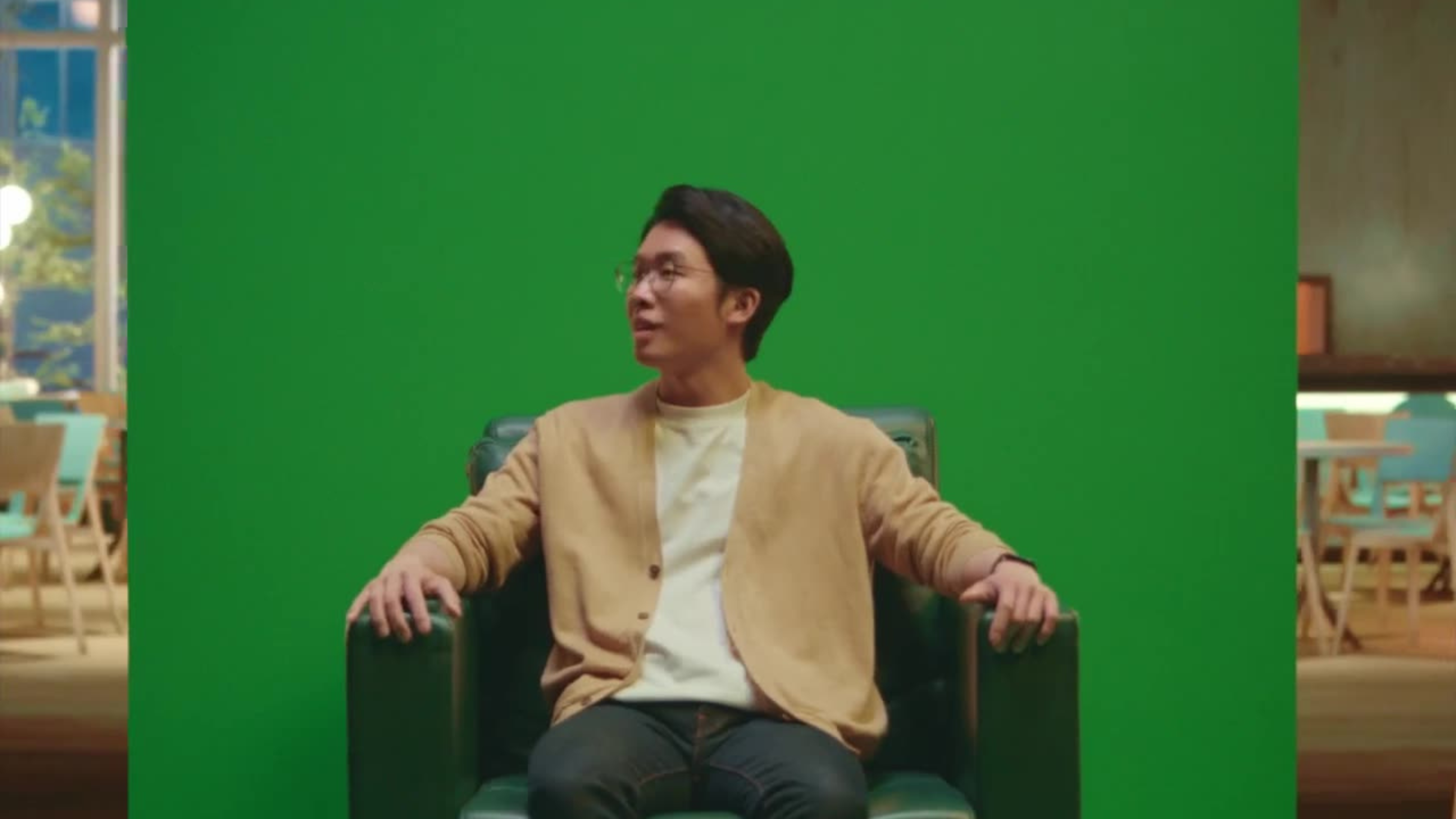 A young man in glasses and a tan cardigan sits in a green armchair in front of a green screen backdrop.
