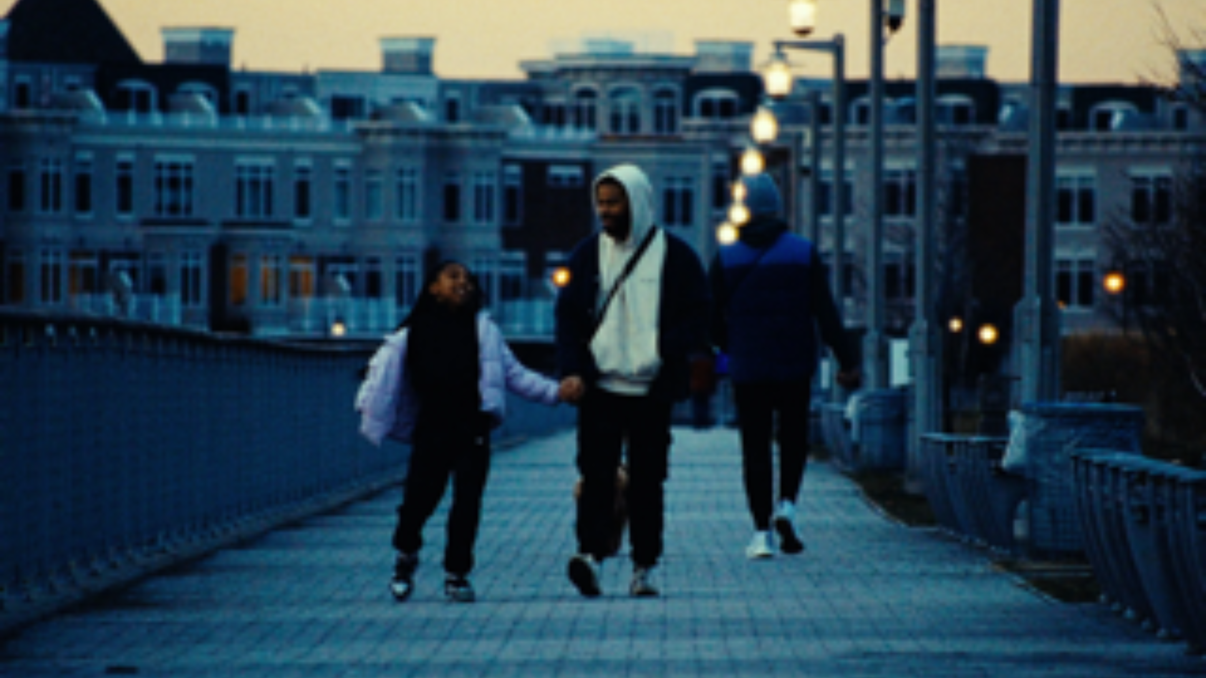 A father and daughter walk hand in hand along a city boardwalk at sunset.