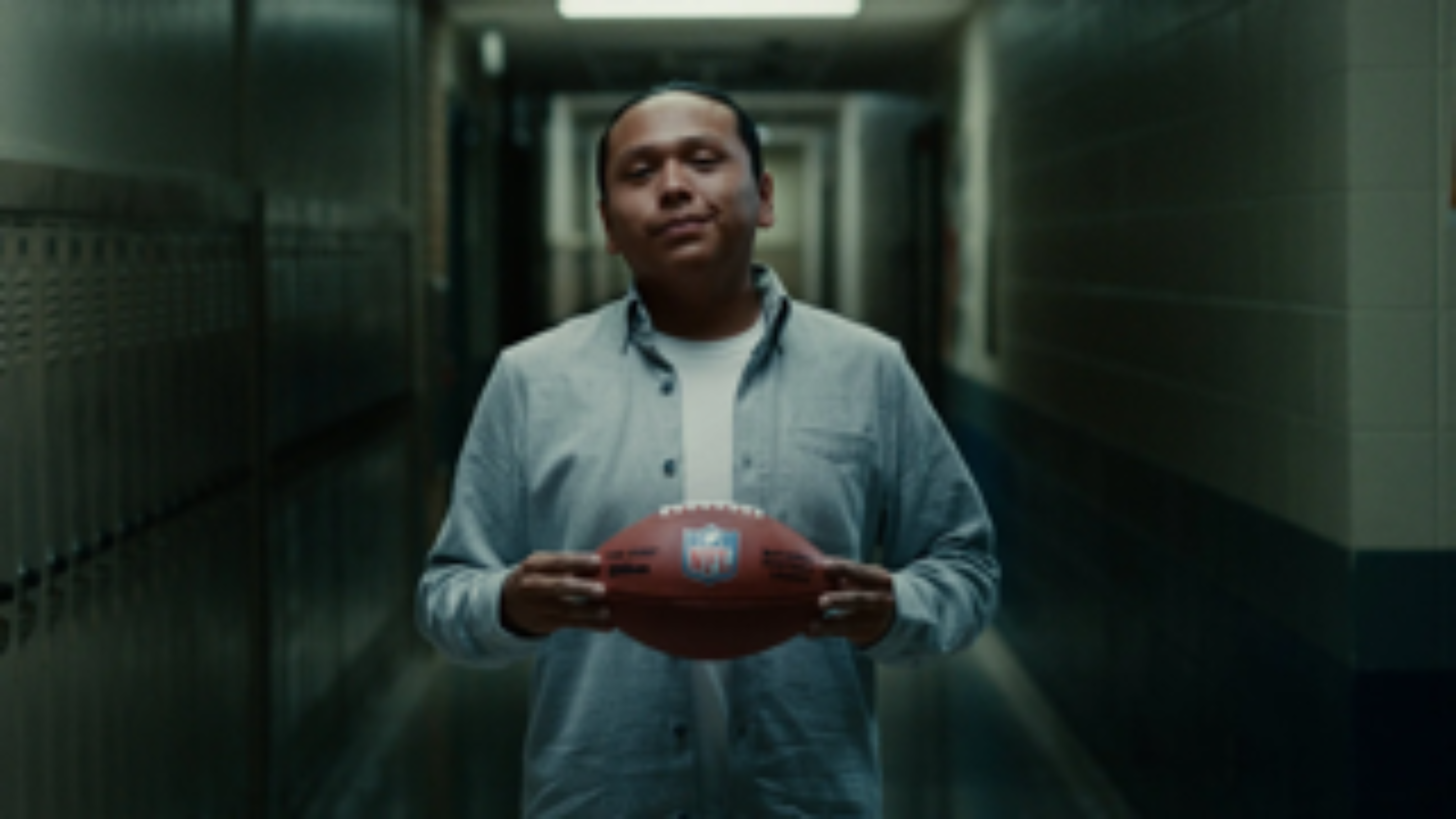 A man stands in a dim hallway holding a football, symbolizing pride and perseverance.