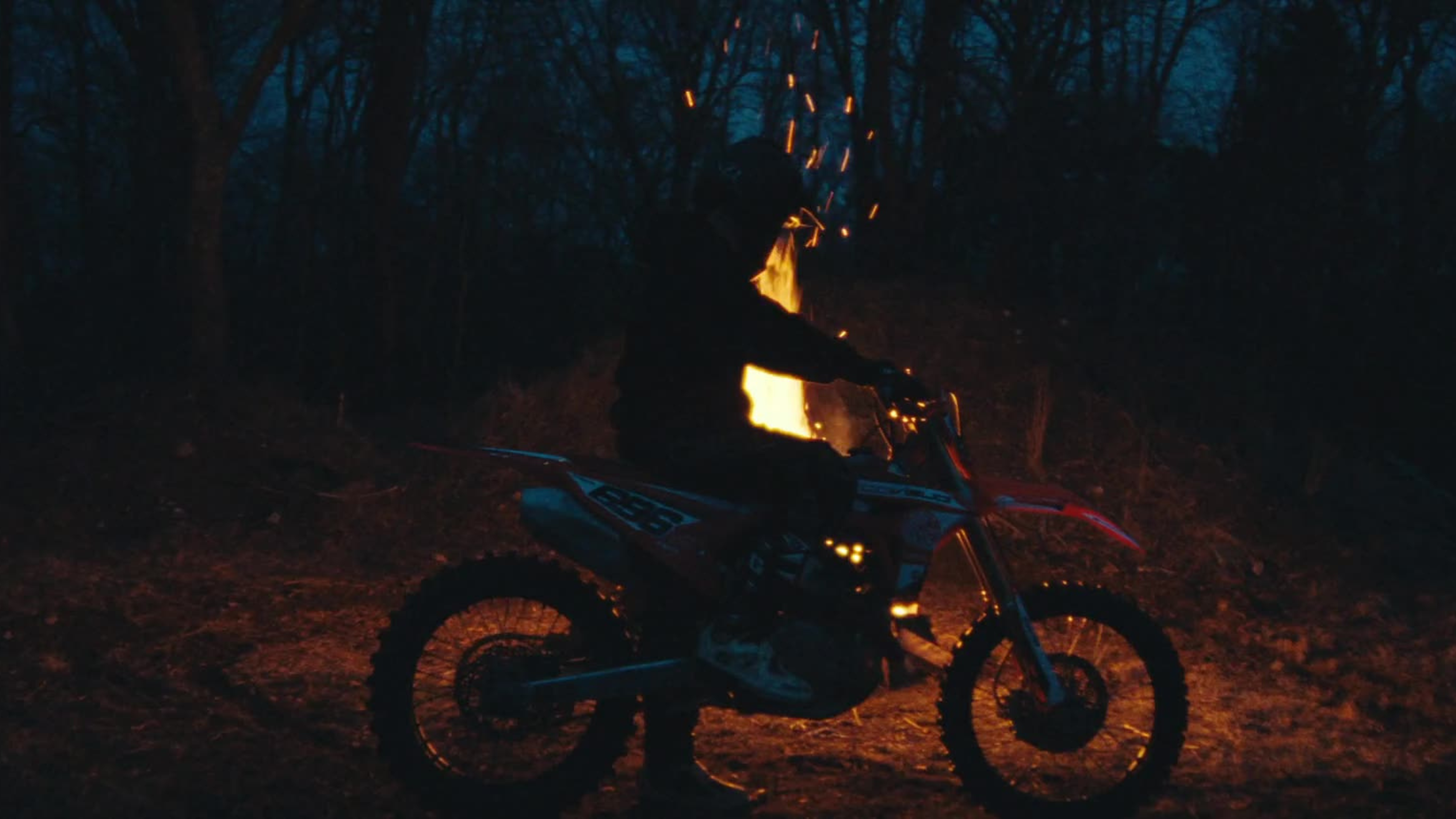 A dirt biker sits near a roaring fire at night, illuminated by sparks and moody forest lighting.