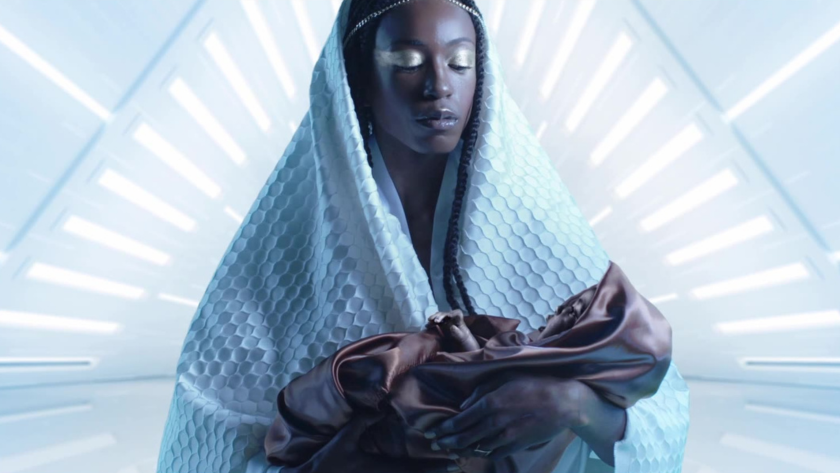 A serene, ethereal woman draped in a white, honeycomb-textured robe cradles a silky bundle as if it were a child. Bathed in white-blue light with radial lines forming a halo effect behind her, this powerful composition merges religious iconography with sci-fi futurism.