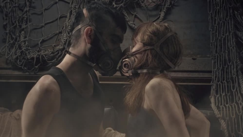 A gritty and emotional moment between two individuals wearing gas masks in a dim, industrial environment. The pair leans in as if to kiss, creating a poignant contrast between survivalist harshness and human vulnerability—conveying themes of love and resilience in a dystopian world.