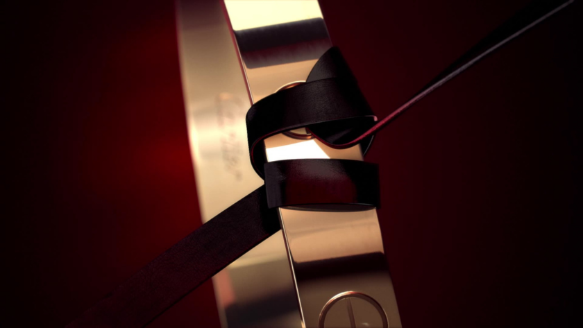 A visually sensual close-up of a gold bracelet cinched with a sleek black ribbon against a deep red background. The soft reflections and sculptural lighting suggest a high-end product reveal—combining precision, allure, and luxury aesthetics often used in jewelry or fragrance campaigns.