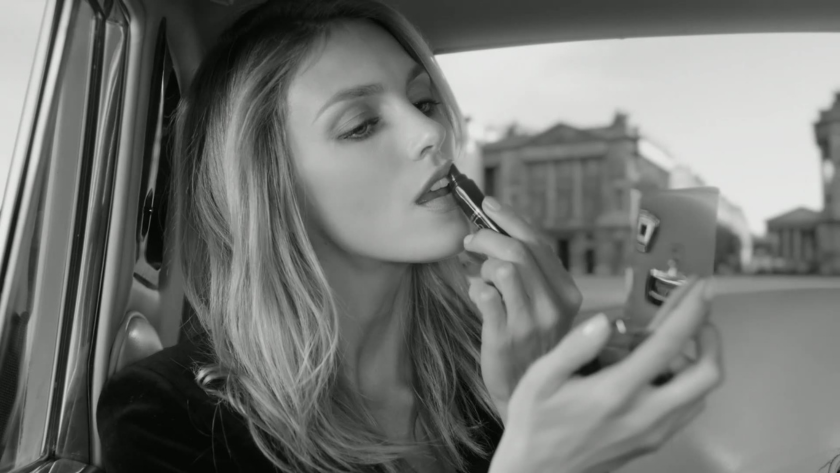 In a timeless black-and-white frame, a woman applies lipstick using a compact mirror while seated in a car, with stately architecture visible through the window. The composition evokes elegance and quiet confidence, capturing a moment of self-styling with fashion editorial finesse.