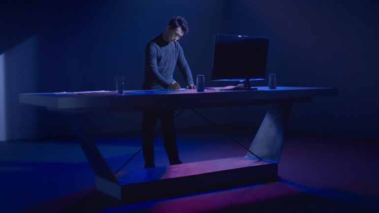 A man stands in a futuristic, dimly lit studio, immersed in his work at a sleek, geometrically designed desk illuminated by a blend of deep blue and magenta lighting. The scene is minimal yet stylized, featuring a curved monitor, glass of water, and modern accessories—conveying themes of innovation, solitude, and focused creativity.
