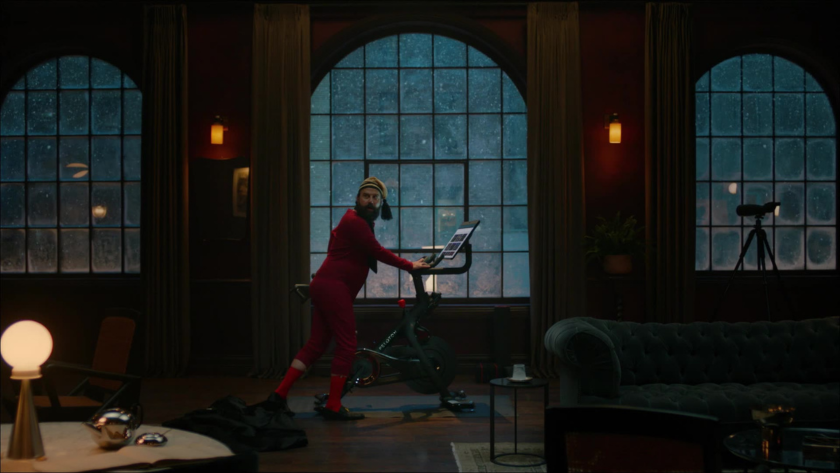 A man in festive pajamas pedals on a Peloton in a moody, snow-lit room, blending comedy with holiday fitness culture.