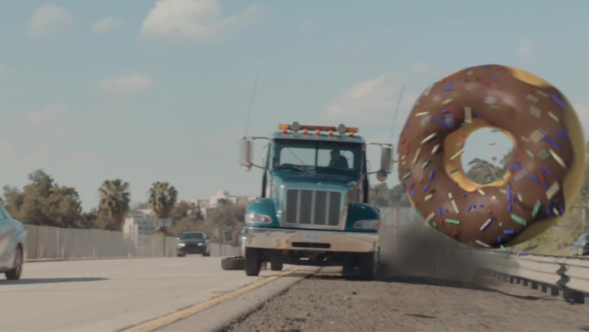 A massive donut crashes down a freeway beside a semi-truck in a high-speed surreal chase, showcasing Toyota’s playful narrative edge.
