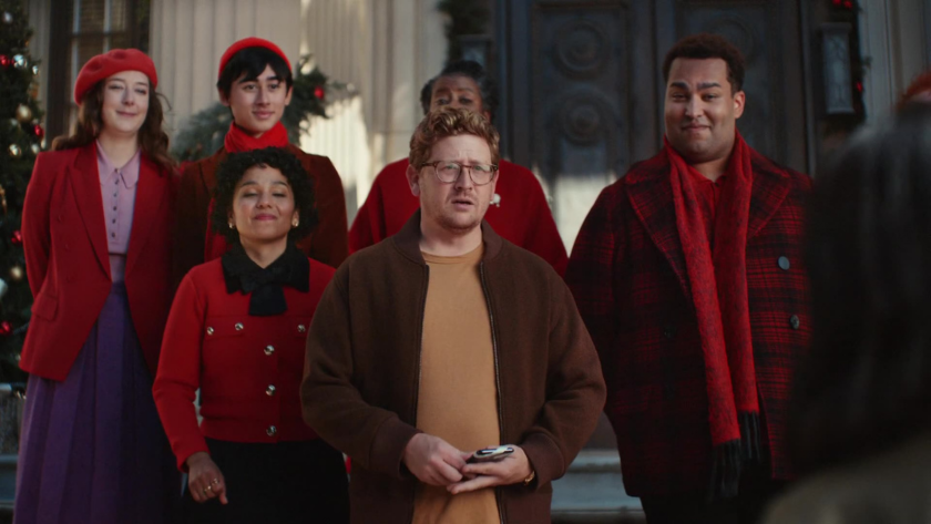 A group of diverse holiday carolers stands on a doorstep, blending tradition with a modern twist on festive connection.