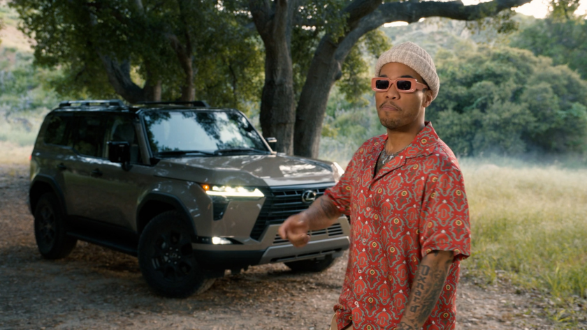 A man in a patterned red shirt and sunglasses stands confidently in front of a parked Lexus SUV beneath the trees.
