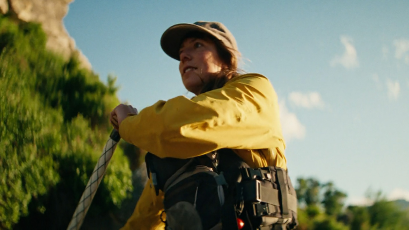 A woman in a yellow jacket grips a rope with determination against a lush, sunlit backdrop.