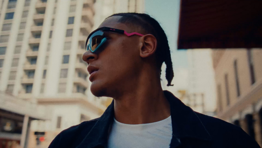 A man in sunglasses and denim glances upward on a city street.