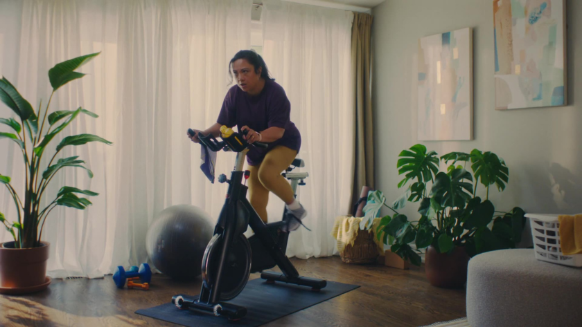 A woman rides a stationary bike in a bright, plant-filled living room, with warm natural light streaming through sheer curtains to energize the frame.