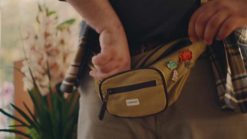 A close-up of a person's hand zipping a mustard-colored fanny pack adorned with enamel pins, framed to emphasize texture and detail in a casual indoor setting.