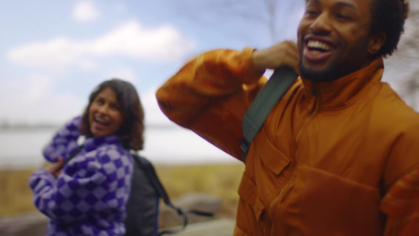 Two hikers laugh together as they sling on backpacks outdoors, captured with a shallow depth of field to spotlight their expressions and movement.