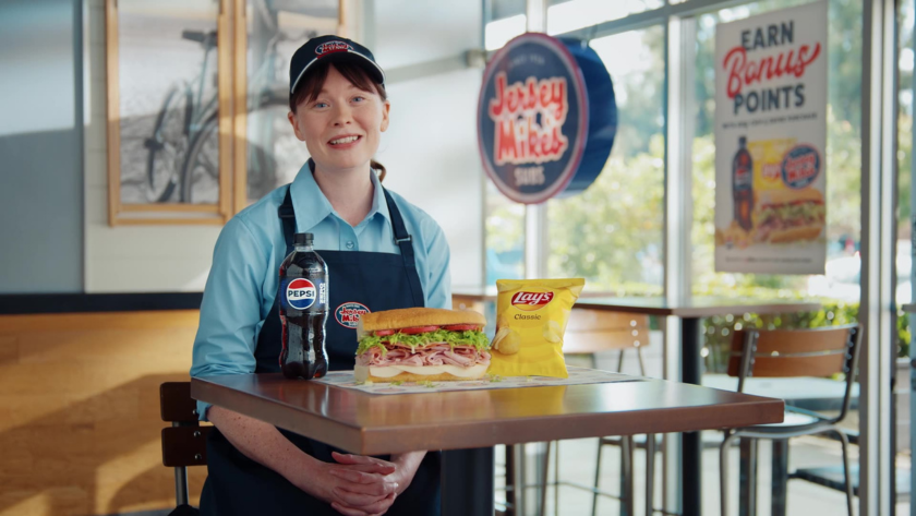 A smiling employee in uniform presents a sandwich, chips, and soda at a restaurant table, framed with bright, even lighting for clear brand visibility.