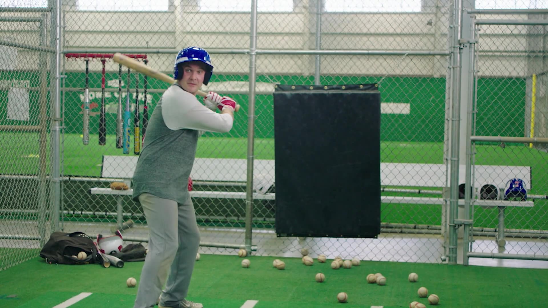 A batter takes a swing in an indoor baseball batting cage strewn with practice balls.