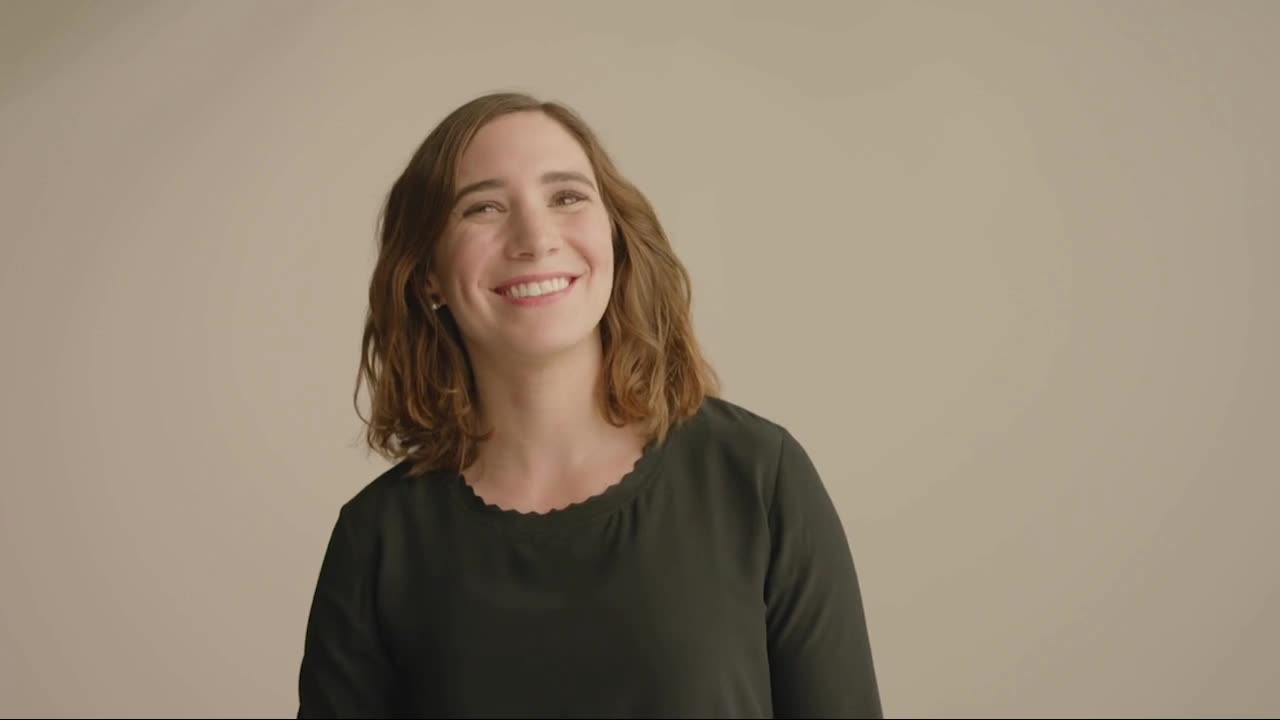 A smiling woman in a dark top laughs naturally against a plain studio background.