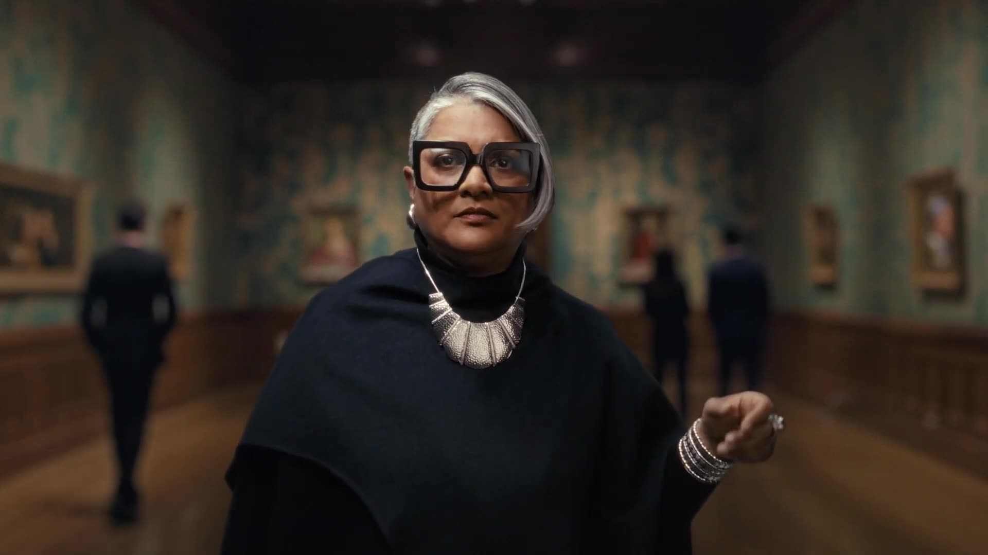 A stylish woman in bold glasses and a statement necklace stands in an art gallery.