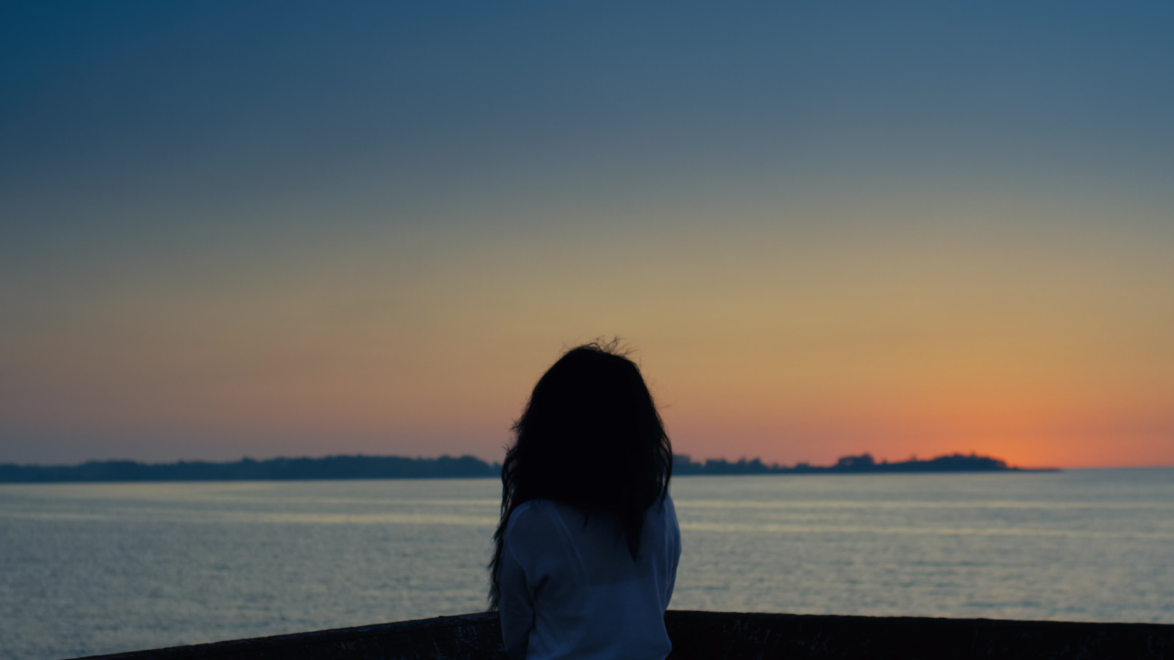 Contemplative silhouette of a woman gazing across water at sunset, captured in serene blue and amber tones.