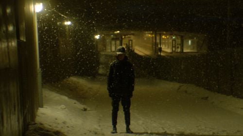Man dressed in winter clothing standing on a sidewalk covered in snow with snow falling. Gloomy setting.