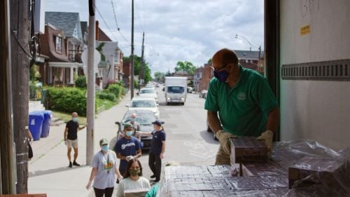 Photo of man giving out boxes from the back of a truck to a line up of people in a home lined street 