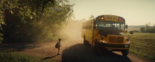  A young boy standing on the side of the road wearing a baseball cap and back-age. A school bus is pulling up on the dirt road beside him. Trees are to the left of the boy and a field is on the other side of the bus.