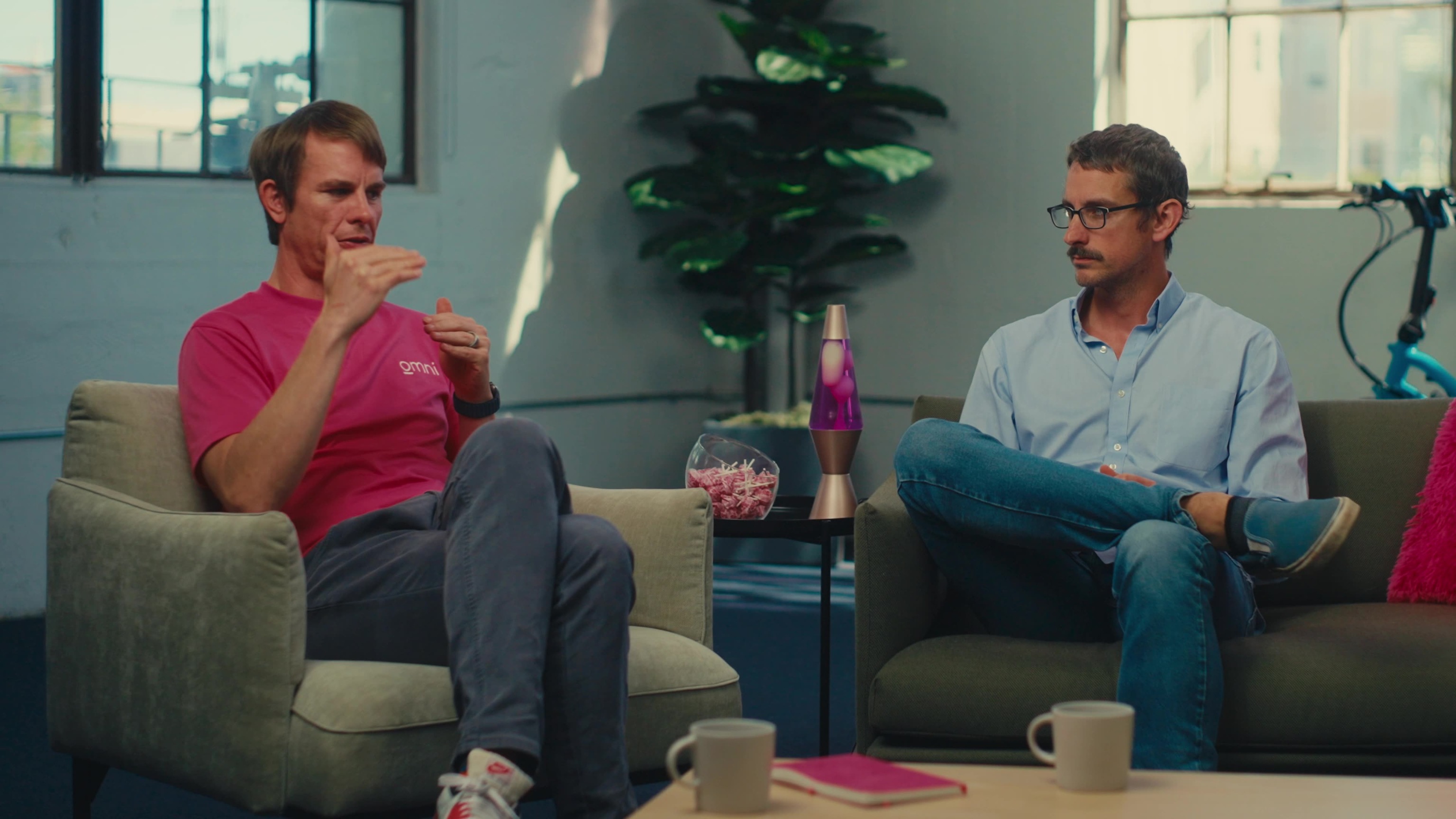 Two Omni team members converse in armchairs across a coffee table, the man in pink mid-gesture explaining a point to his colleague in blue.