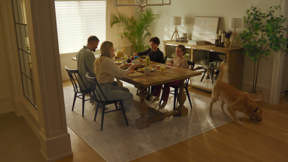A family sits around a wooden dining table sharing a meal in a bright, cozy home while a dog walks through the room beside them.