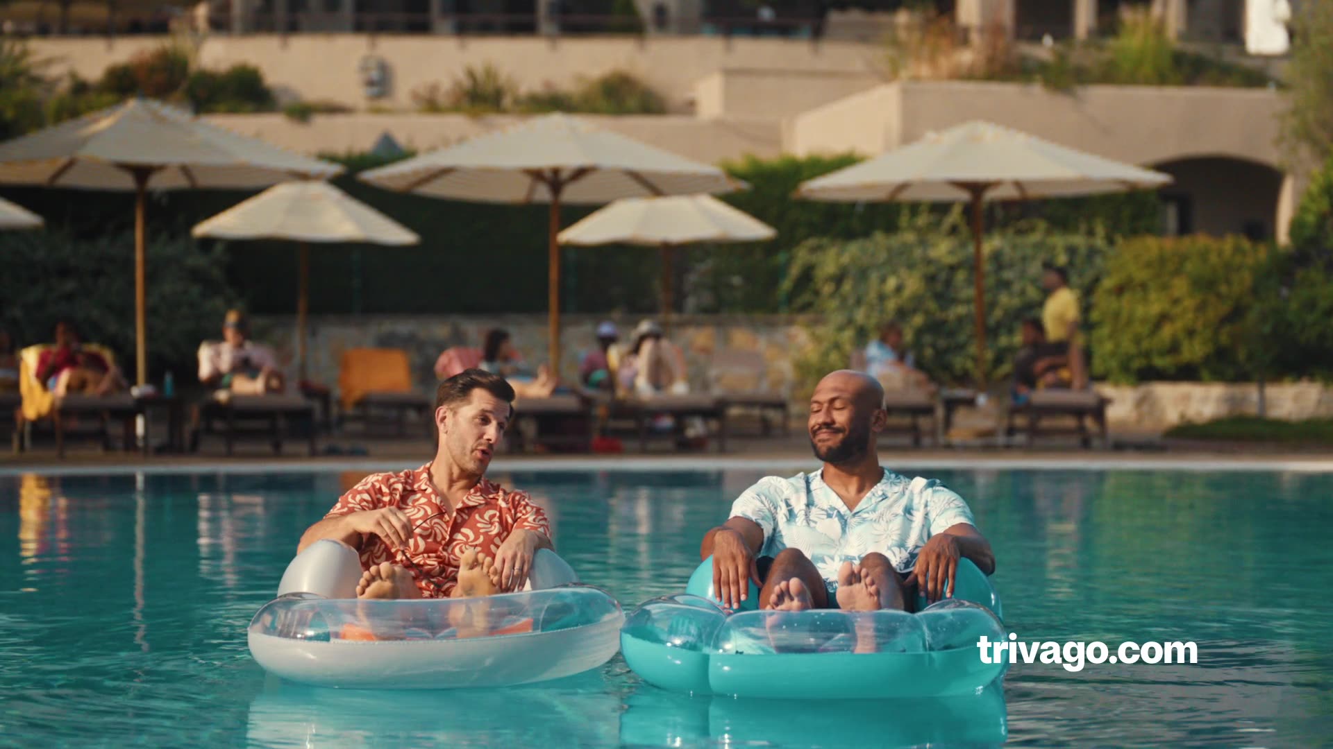 Two friends relax and laugh together in a resort pool at golden hour with palm trees and buildings behind them.