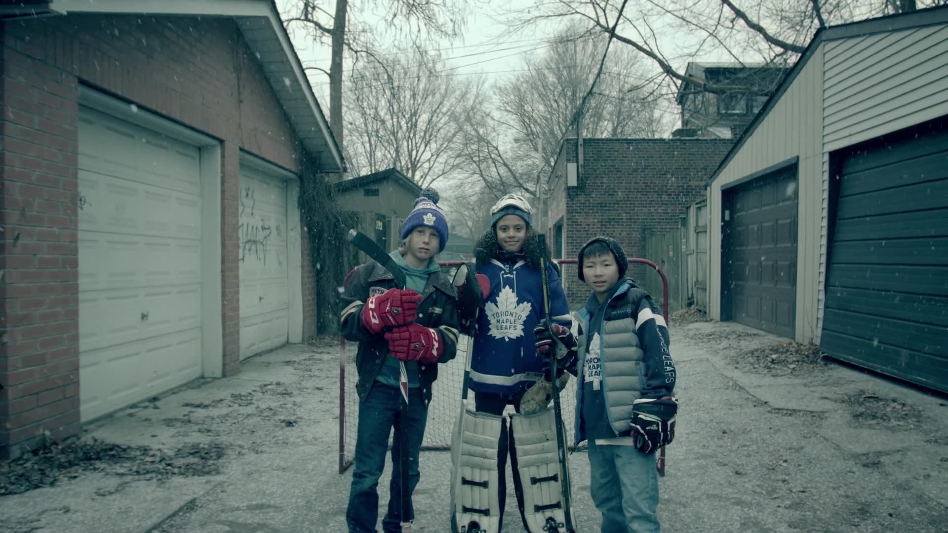 Three young Toronto Maple Leafs fans in jerseys and hockey gear pose with sticks and a net in a snowy Toronto alleyway.
