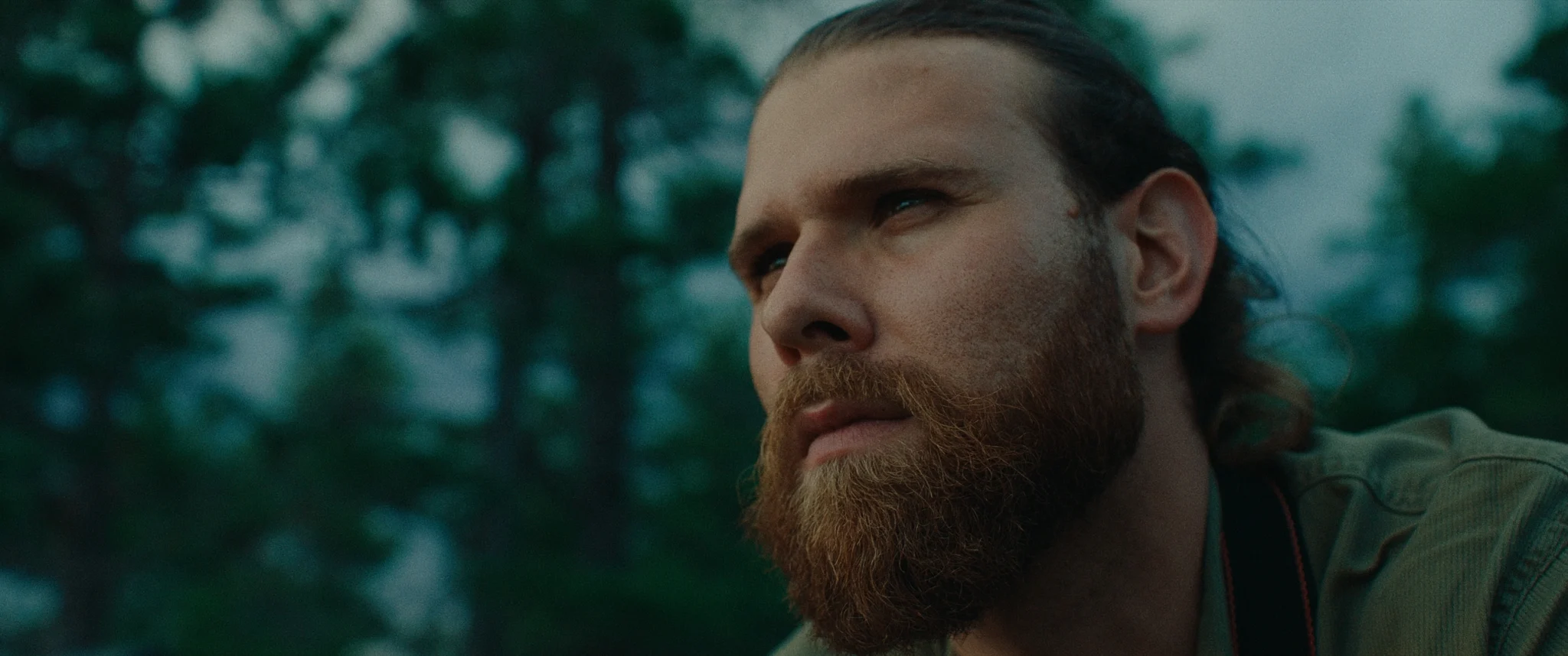 Man with beard and pony tail looking straight ahead with trees in background