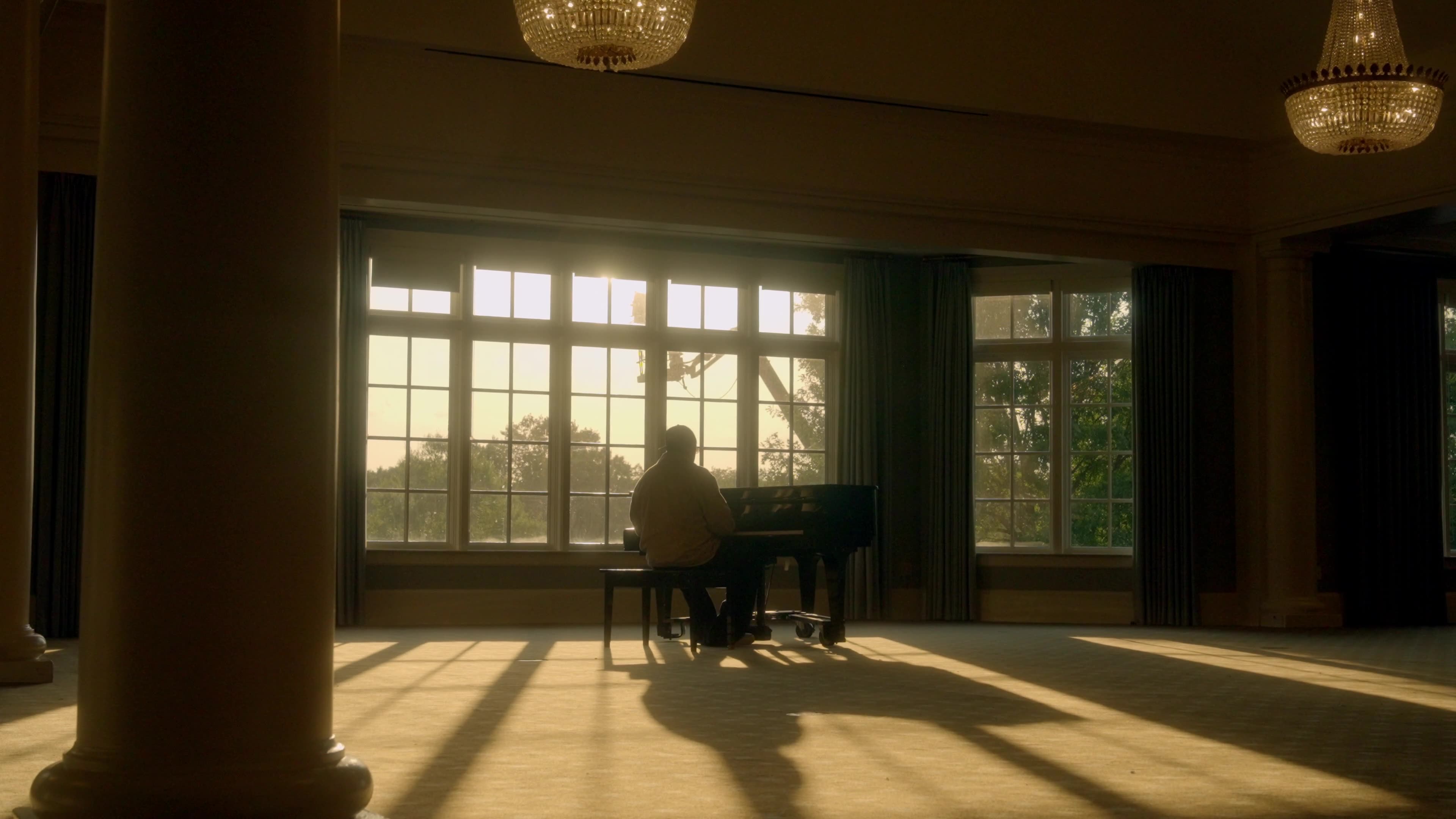 A lone figure sits at a piano in a grand sunlit room with tall windows
