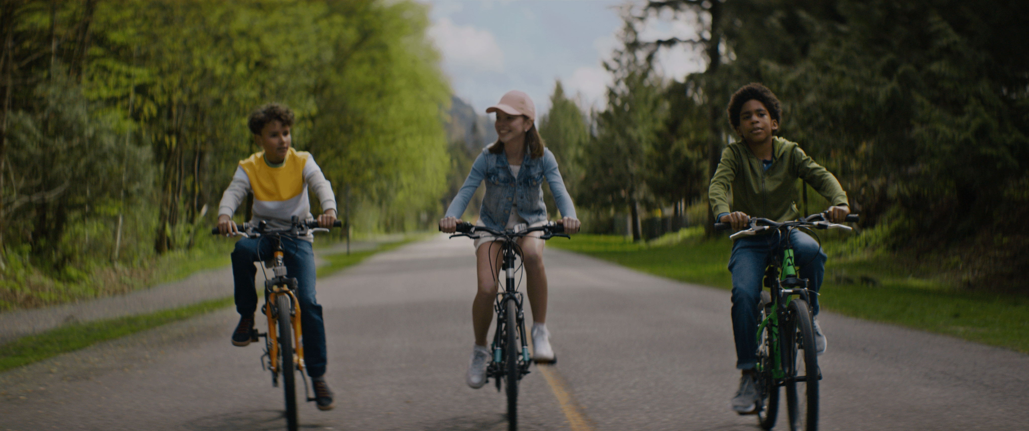 Three kids on their bikes riding in the middle of an empty road with trees on both sides of them. 